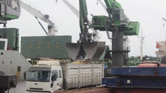 A large green loader with a Rotobec clam bucket in a seaport filling a truck with sand from a ship.